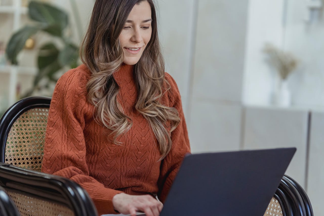 Crop smiling female in red sweater using modern netbook while sitting on cozy armchair in light living room